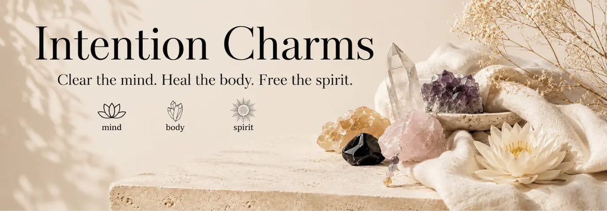 A styled promotional graphic for 'Intention Charms' with crystals, dried flowers, and a stone slab on a warm beige background. Text reads 'Clear the mind, heal the body, free the spirit".