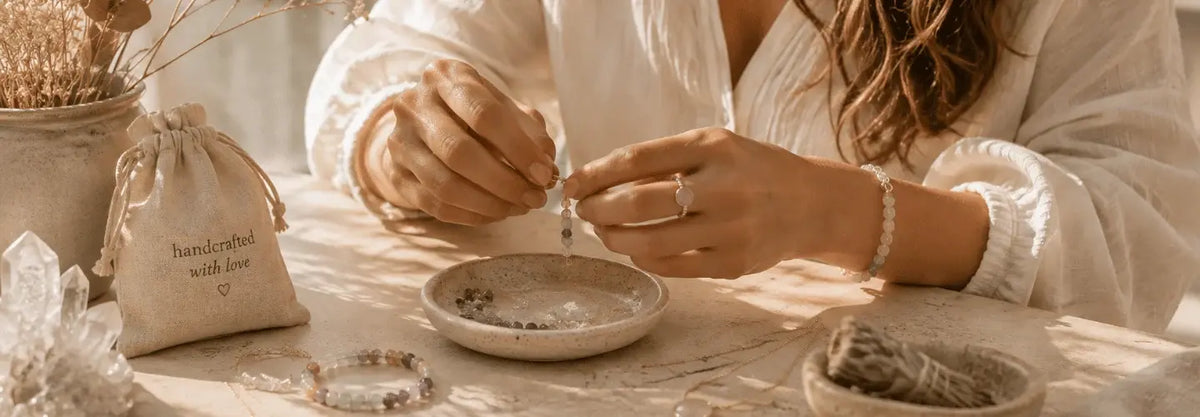 A close-up lifestyle shot of a person's hands crafting a bracelet in a warm, sunlit boho setting. The person is wearing a white blouse and a delicate bracelet, with a small beige drawstring bag printed with 'handmade with love' on the table alongside crystals and sage.