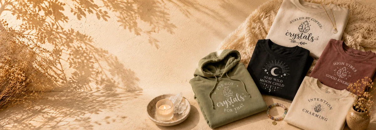 Shop clothing banner for Intention Charms featuring a styled flat-lay of four hoodies and t-shirts in earthy tones, featuring celestial and crystal designs. The garments are arranged on a textured beige surface with warm tree shadows, alongside a small candle, dried flowers, and a decorative bowl.