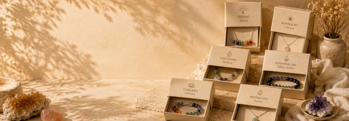 Shop by product line banner for intention charms featuring several open jewelry gift boxes arranged on a warm beige surface, showing colorful beaded bracelets and necklaces inside. The scene is styled with crystals, a candle, dried flowers, and soft shadow overlays on a beige background.